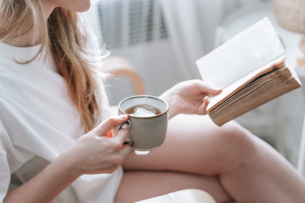 Crop anonymous female with crossed legs and cup of hot drink reading aged textbook in armchair at home